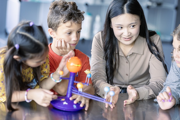 A small group of elementary students gather around a table with their science teacher as they explore the solar system together. The teacher has a small model of the solar system out in front of them as she teaches the lesson.