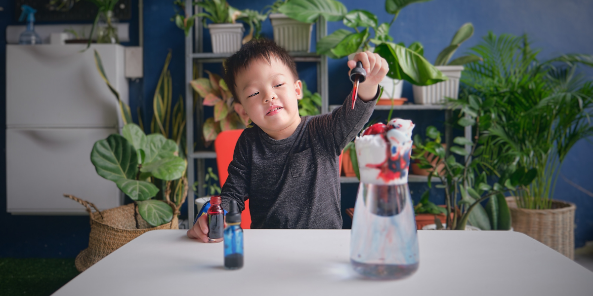 Young child in dark sweater using red pipette during STEM-Inspired Kitchen Table Project, transferring colored liquid into glass container for density tower experiment, showcasing hands-on learning and fine motor skills
