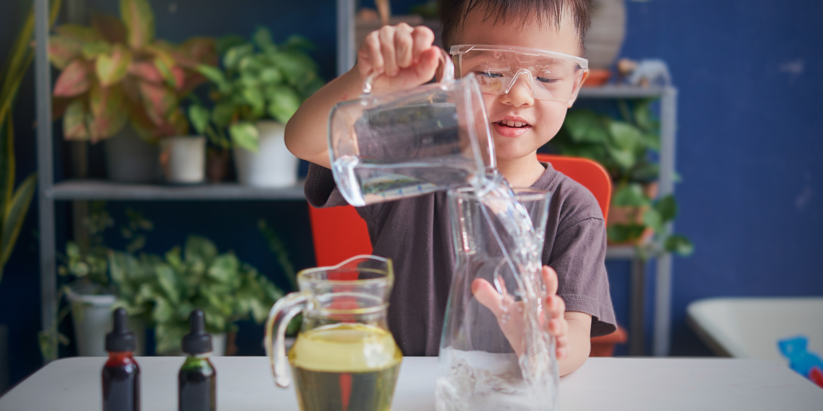 Young child wearing clear safety goggles carefully pouring water during STEM-Inspired Kitchen Table Project, demonstrating proper lab safety practices in home-based density science experiment for elementary students