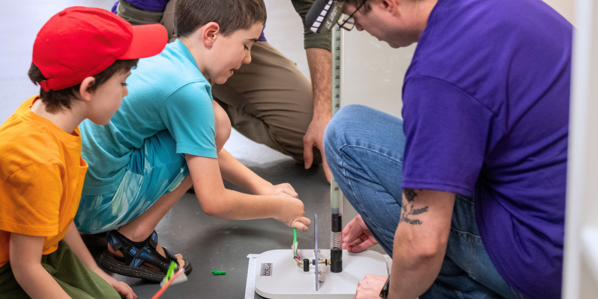 Students and instructor examining engineering equipment together during a hands-on STEM lesson, demonstrating the mentorship approach of Engineering For Kids<sup srcset=