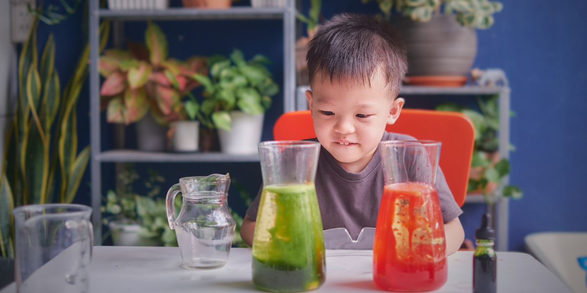 Child observing successful STEM-Inspired Kitchen Table Project results with separated red and green liquid layers in glass containers, showcasing completed Valentine's Day themed density experiment on white table