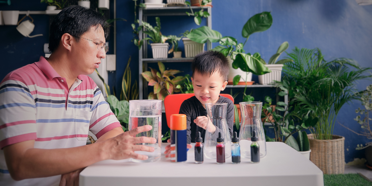 Parent supervising young child during STEM-Inspired Kitchen Table Project featuring colorful liquid density experiment on white table surrounded by houseplants, perfect Valentine's Day science activity for kids at home
