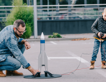 Father and young boy getting excited about space science while launching a model rocket in a parking lot, demonstrating hands-on learning about aerospace principles.