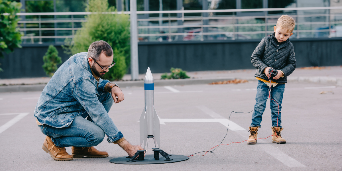 Father and young boy getting excited about space science while launching a model rocket in a parking lot, demonstrating hands-on learning about aerospace principles.