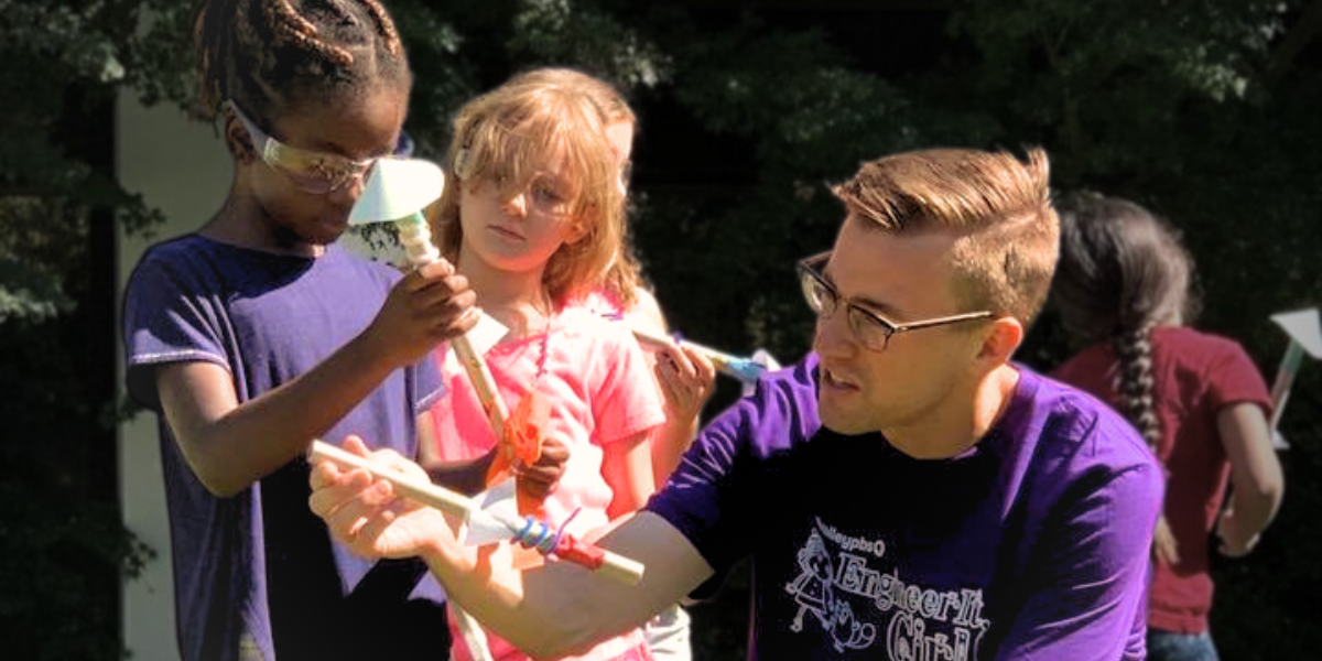 Space camp instructor helping diverse children build a model rocket, fostering excitement about space engineering through practical outdoor activities.